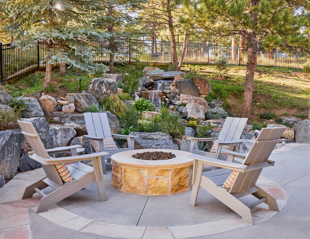 Outdoor seating area with four gray chairs around a stone fire pit and landscaped rocks.