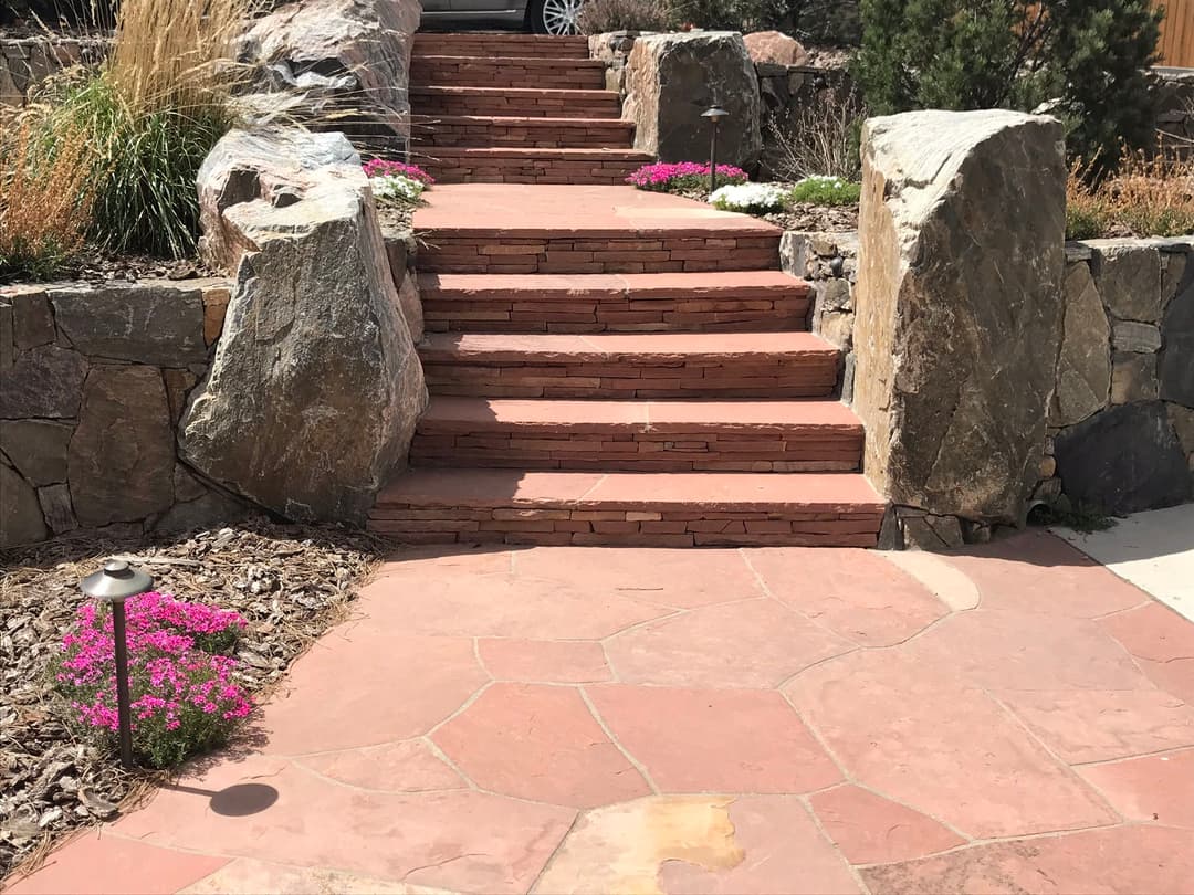 Stone steps with pink flowers lead to a home, bordered by large rocks and decorative lighting.