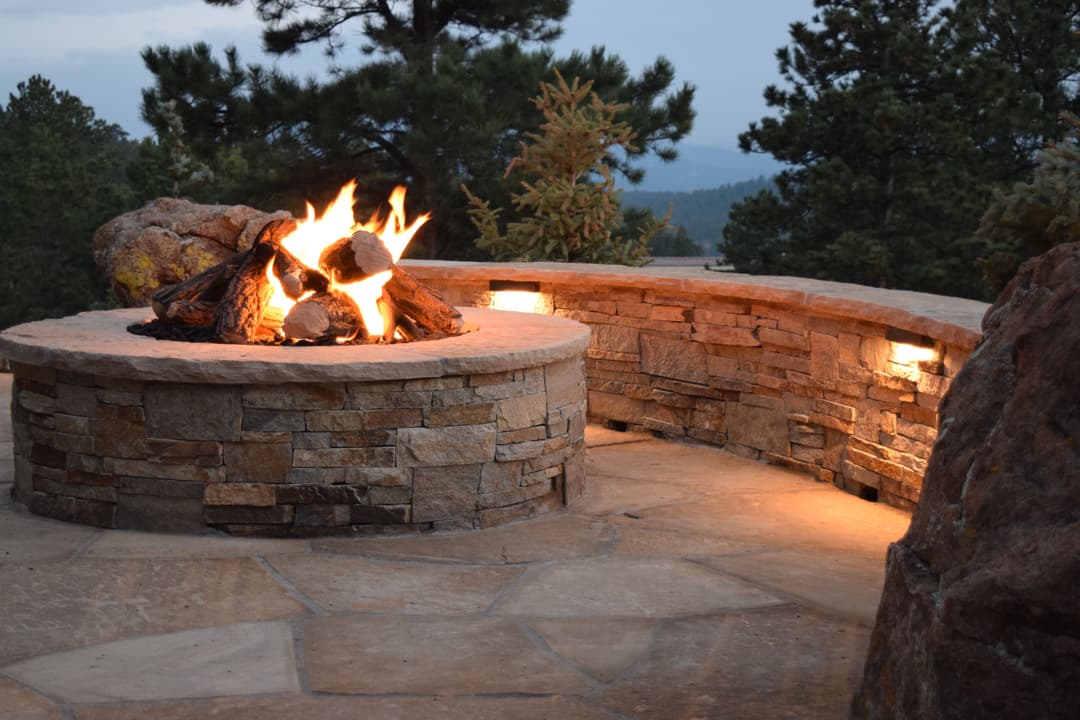 Stone fire pit with flames, surrounded by trees and mountains at dusk. Cozy outdoor setting.