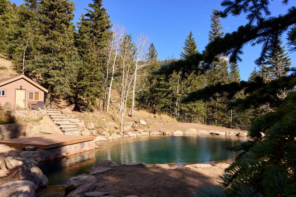 Serene natural hot springs surrounded by pine trees and a wooden cabin in a mountainous setting.