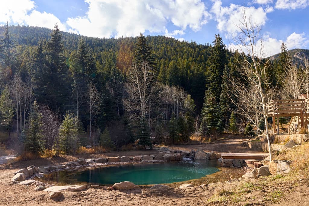 Natural hot spring surrounded by evergreen trees and mountains under a blue sky.