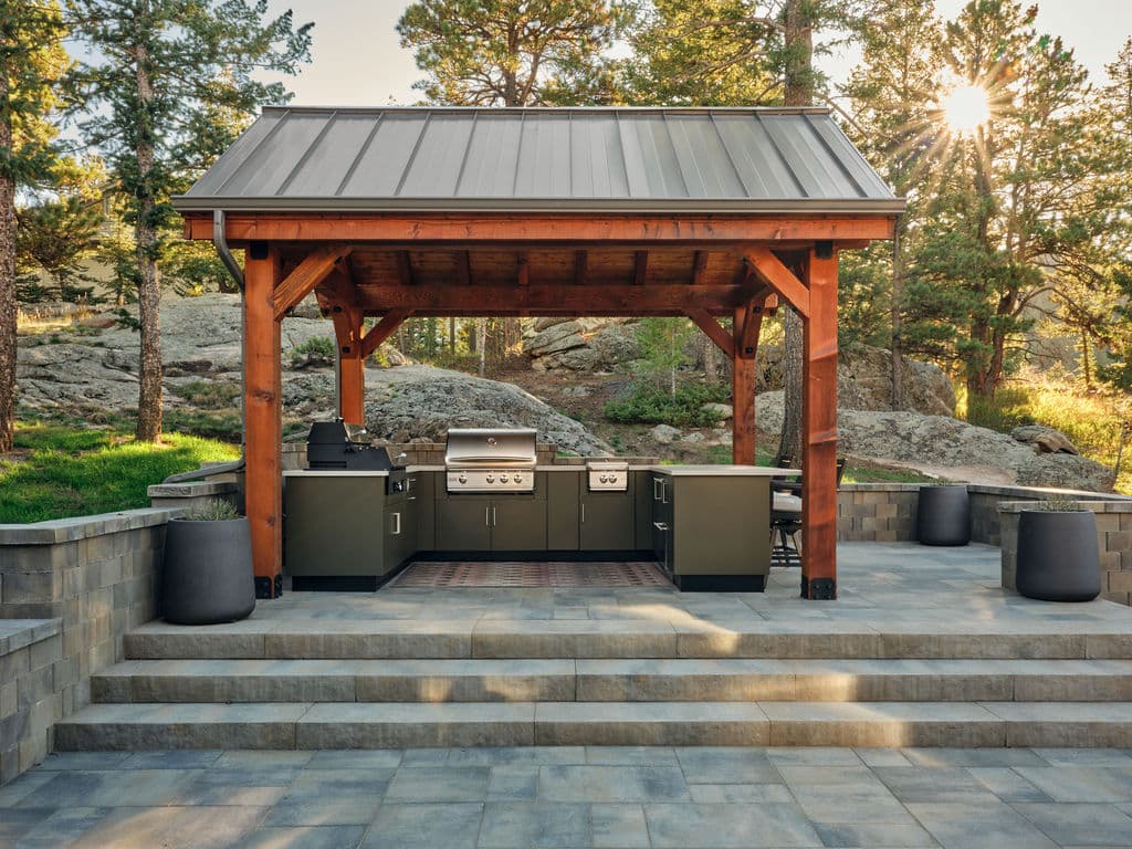 Modern outdoor kitchen under wooden pavilion with grilling area and stone patio. Sunlight shines through trees.