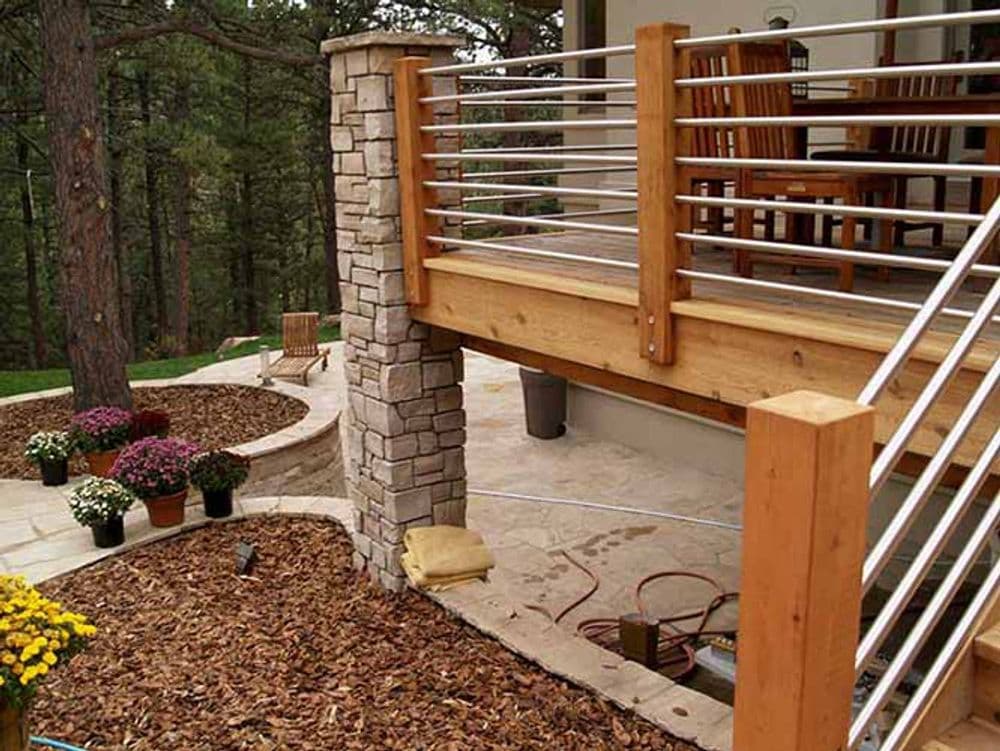 Wooden deck with stone columns, stainless steel railing, and landscaped garden featuring flowers.