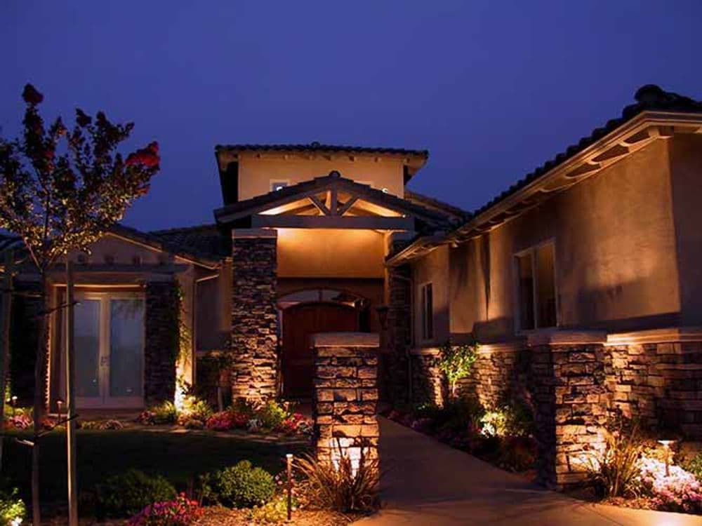Modern house exterior illuminated at night with landscaped garden and stone accents.