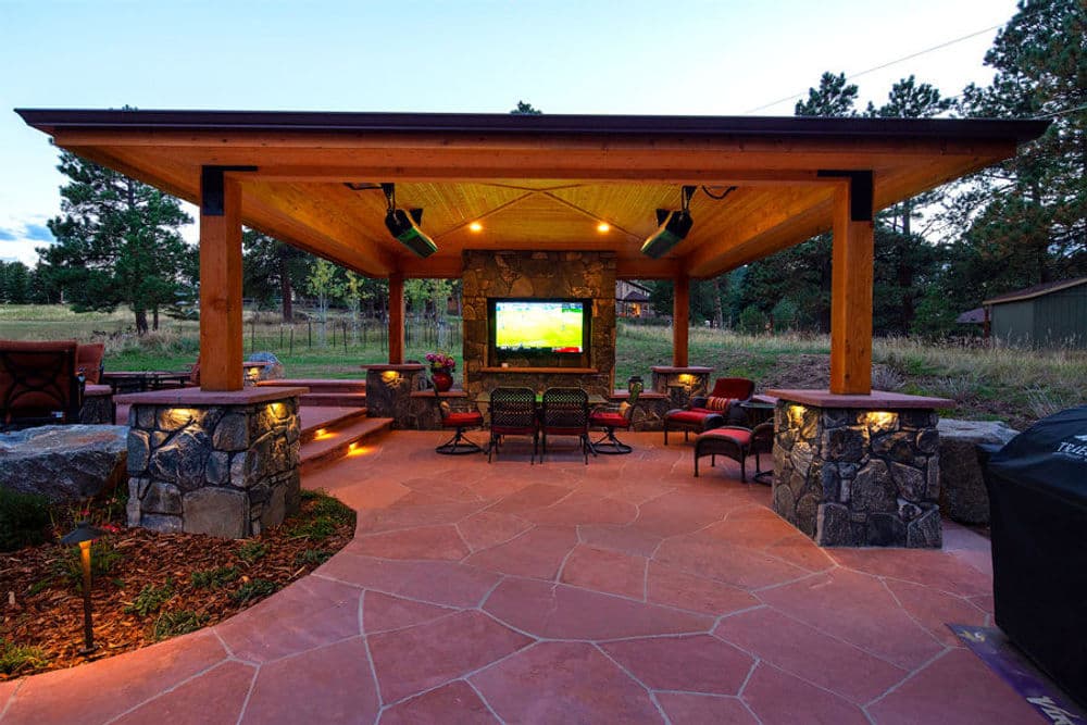 Outdoor patio with stone accents, seating area, and mounted TV under wooden gazebo.
