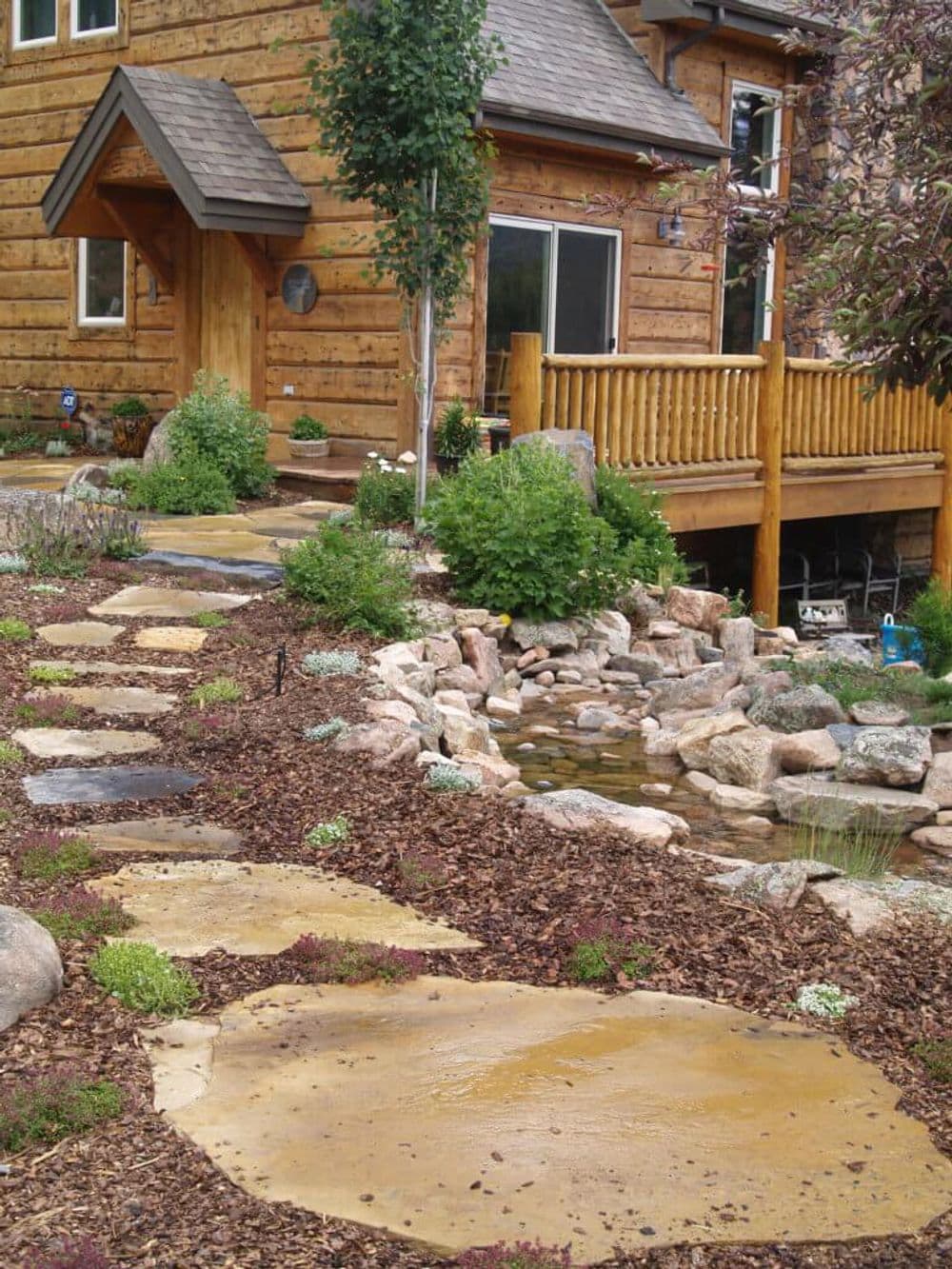 Natural stone pathway leading to a wooden deck, surrounded by lush landscaping and a pond.