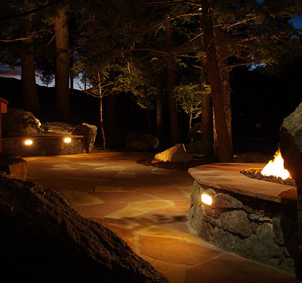 Serene nighttime outdoor patio with stone pathways, fire pit, and soft lighting among trees.