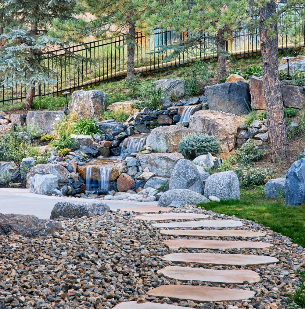 Serene garden with a stone pathway, water feature, and lush greenery among boulders.
