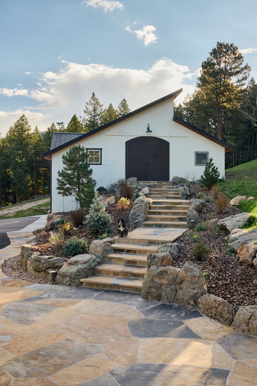 Charming hillside barn with stone steps and landscaping, surrounded by pine trees.
