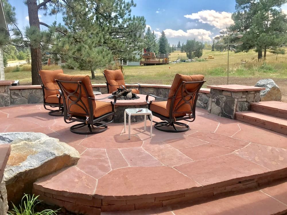 Outdoor patio with four cushioned chairs around a stone fire pit, surrounded by trees and hills.