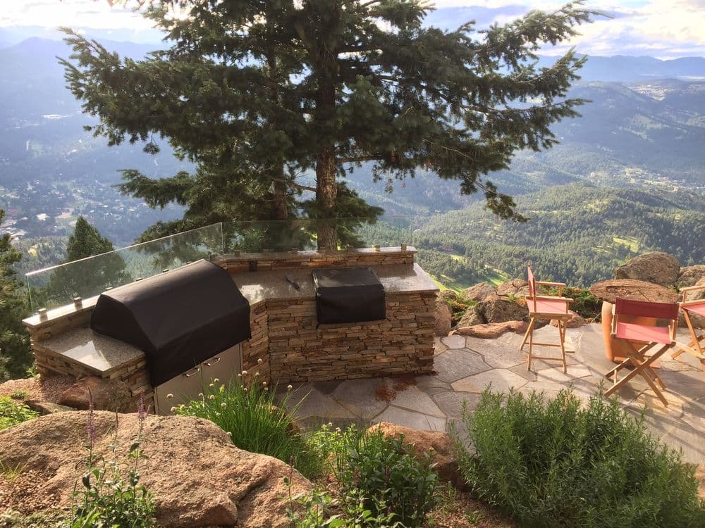 Outdoor kitchen with grill, stunning mountain view, surrounded by natural stone and greenery.